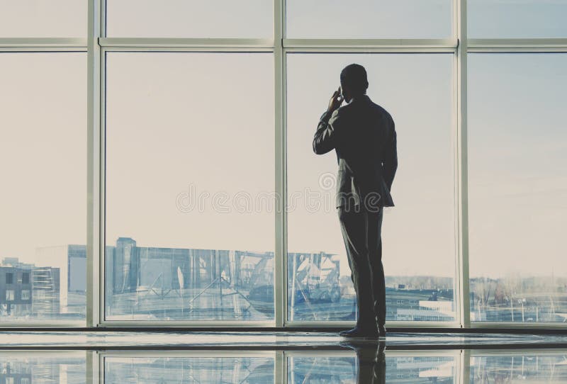 Back view of young businessman is looking out of a panoramic window and talking by phone.  stock photo