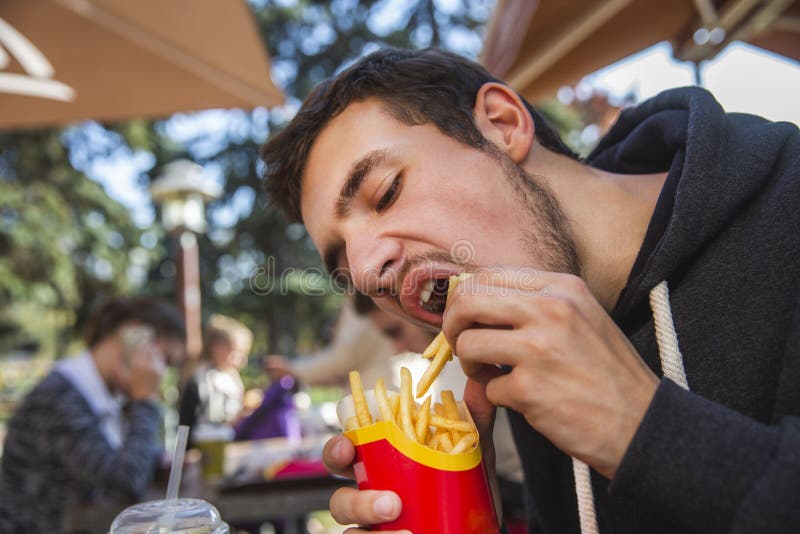 Hungry young man is greedily eating his french fries. He is sitting at the restauraunt terrace.  stock photography
