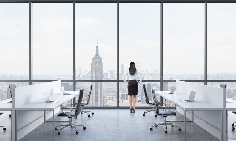 Rear view of a brunette who is looking out the window in the modern panoramic office with New York view. White tables equipped wit. H modern laptops and black stock photo