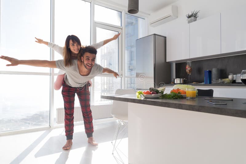 Young Couple In Kitchen, Lovers Hispanic Man Carry Asian Woman Modern Apartment. With Big Windows Interior royalty free stock photography